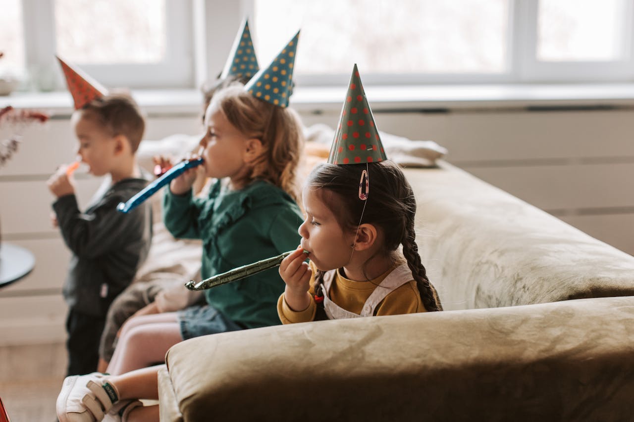 Three young children with party hats enjoy a fun indoor birthday celebration.